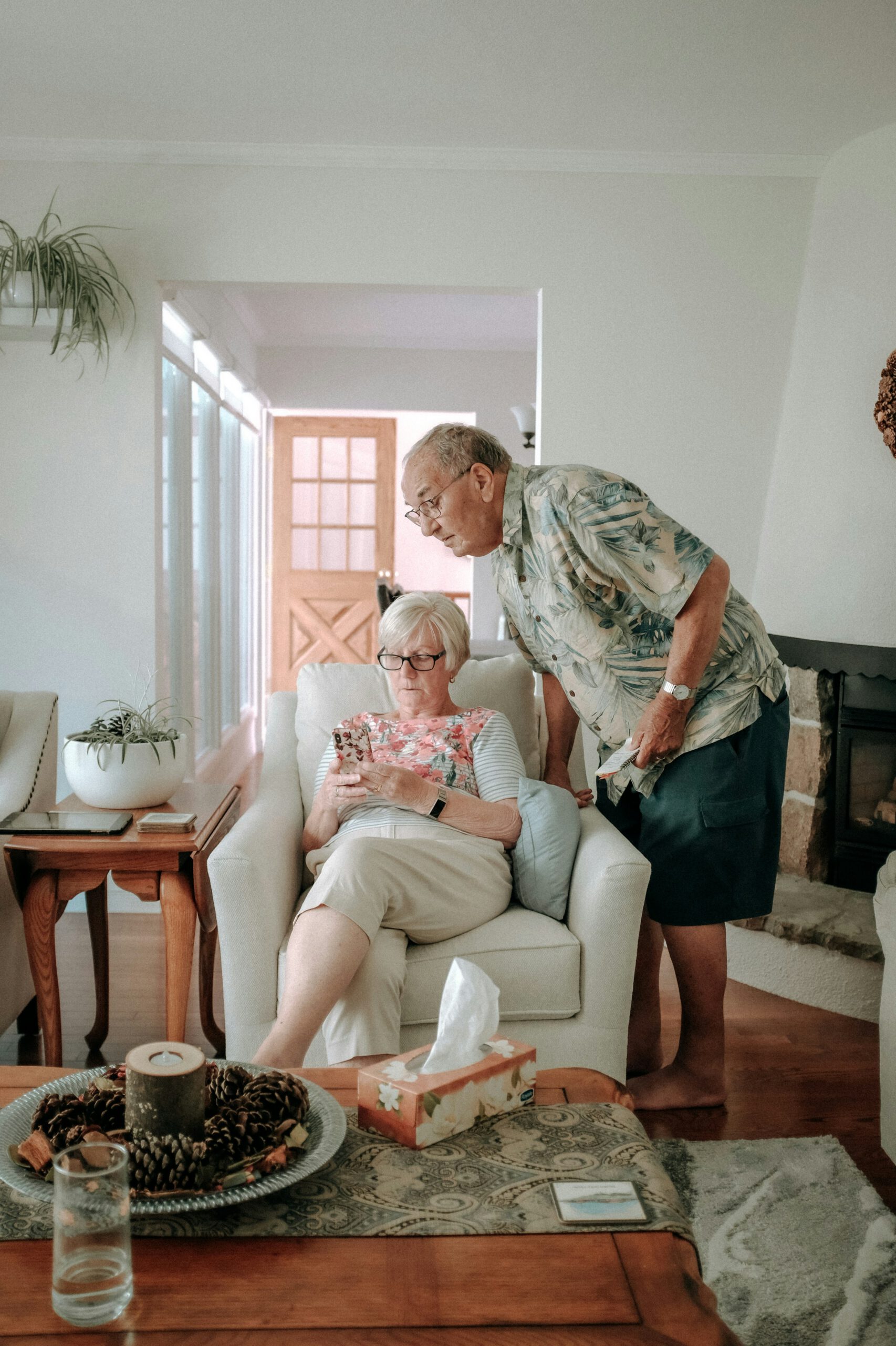 Ein älteres Paar in einem hellen Wohnzimmer: Die Frau sitzt in einem Sessel und schaut auf ihr Smartphone, während der Mann sich über ihre Schulter beugt, um das Display mitzuverfolgen.