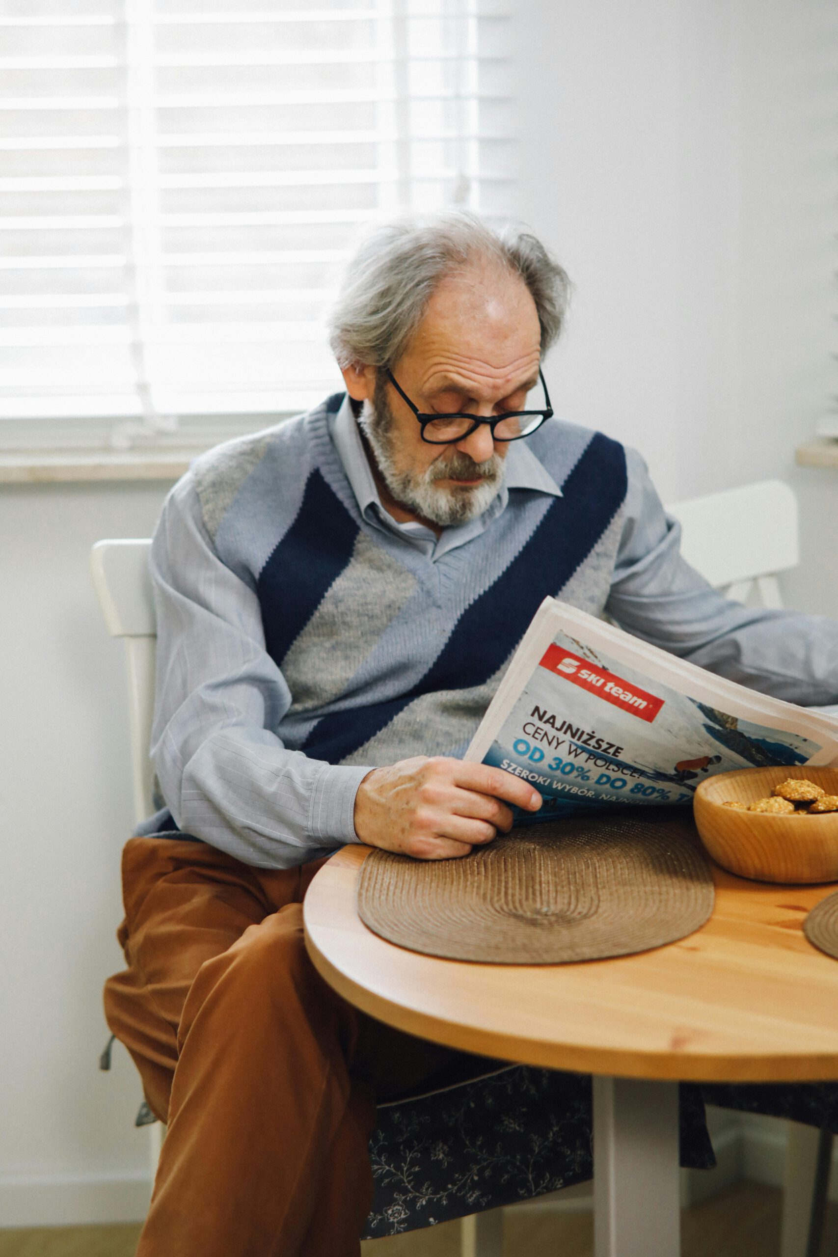 Ein älterer Mann sitzt an einem runden Holztisch und liest konzentriert in einer Zeitung. Er trägt eine Brille sowie einen Pullover mit Rautenmuster und wirkt ganz in seine Lektüre vertieft. Neben ihm steht eine kleine Schale mit Gebäck.